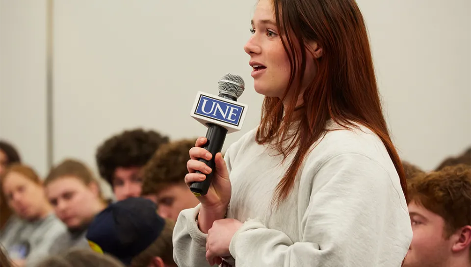 A student speaks into a U N E-branded microphone at a President's Forum event
