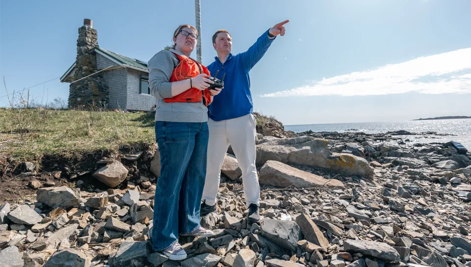 Two U N E student researchers stand on a rocky coastline conducting fieldwork, with one person holding scientific equipment while the other points toward the ocean. Behind them is a historic stone lighthouse building, and the scene shows a rugged shoreline with scattered rocks leading to the water under a partly cloudy sky.