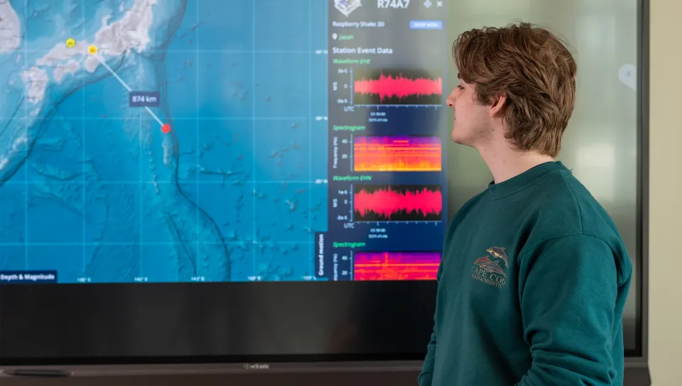 A University of New England student in a green sweatshirt stands in the Sustainable Innovation Center, viewing a large interactive display screen showing seismic data visualization with a map featuring measurement points and colorful spectrograms displaying station event data and waveform analysis.