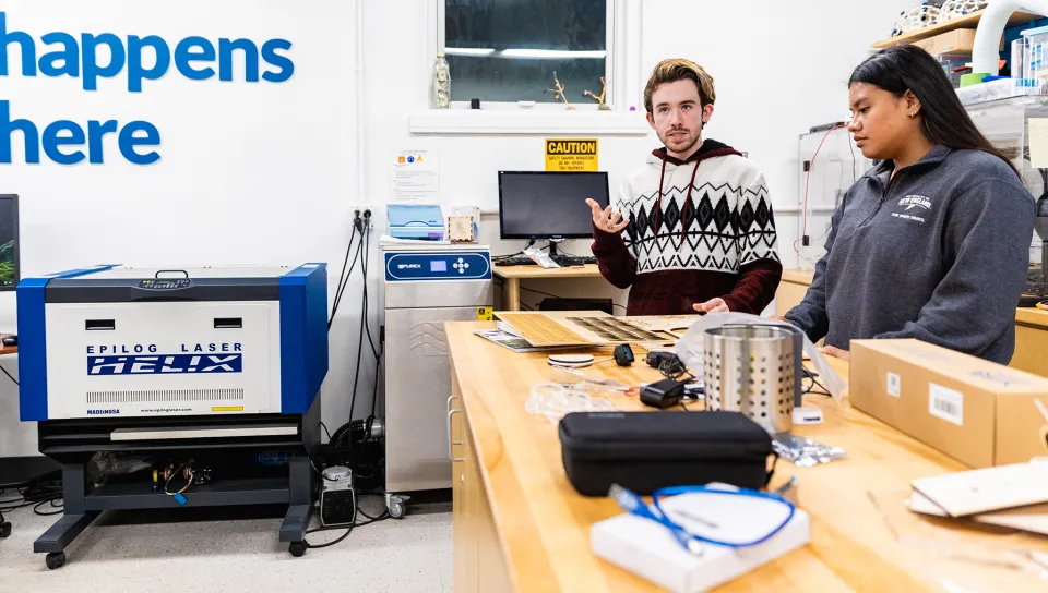 Two students at a long table stand near a laser cutter as they discuss their project