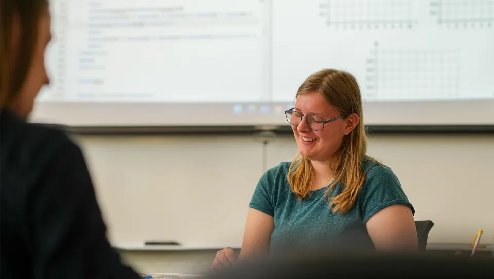 A student wearing glasses smiles during a classroom discussion, with a whiteboard visible in the background.