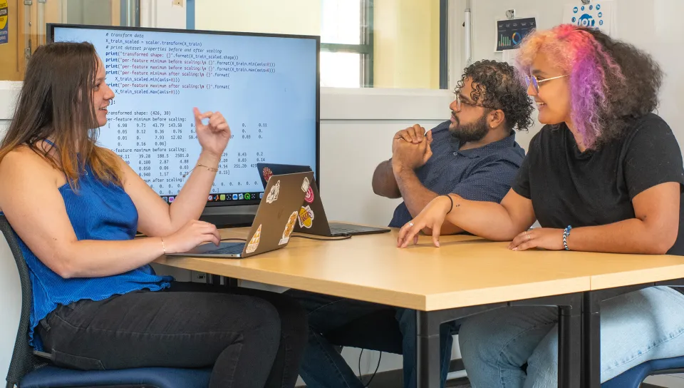 Three students collaborate around a table with laptops, discussing Python code displayed on a large monitor showing data transformation results.