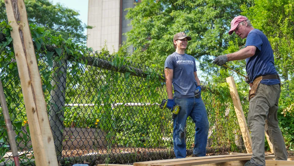Luke Jenkins (Biology, ’26) and Cameron Wake, Ph.D. using gardening tools at a community garden in Portland, Maine, with greenery and campus buildings visible in the background.