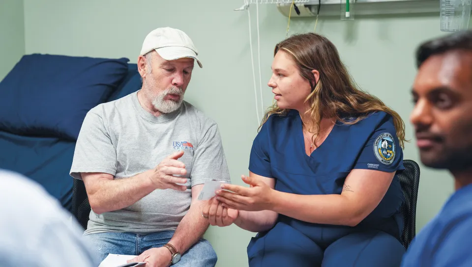 Alexa Lanteri, a nursing student speaks closely with Michael, a patient actor, and hands him a plan sheet while other interprofessional healthcare students observe during the simulation.