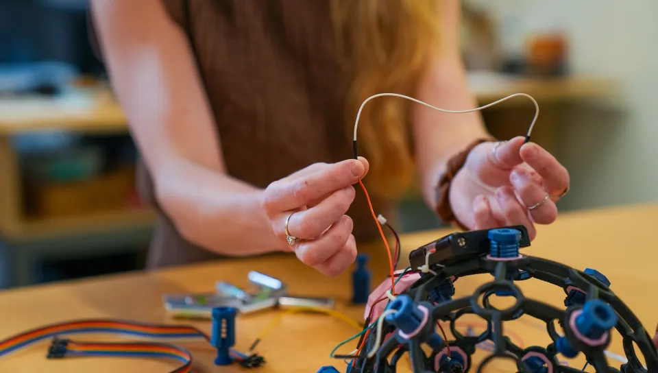 Close-up of hands connecting wire to dome-shaped black EEG sensor array with colored components.