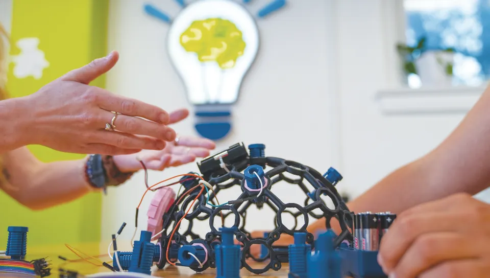 Milo Lypps and Jessica Howard work together on wiring a skull cap with a lightbulb logo visible on the wall behind them.