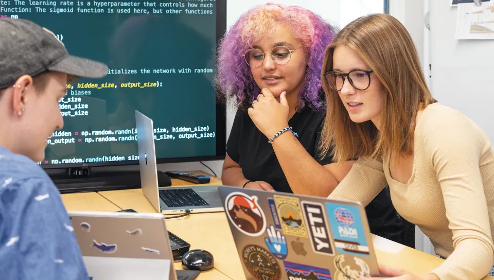 Three students gather around laptops displaying code on a large monitor, engaged in collaborative programming work.
