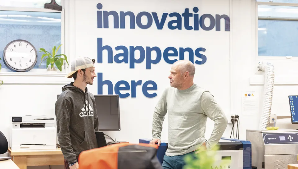 Norm O'Reilly and a studen converse in front of a wall featuring the text "innovation happens here" in a makerspace equipped with laser cutters and 3D printers.