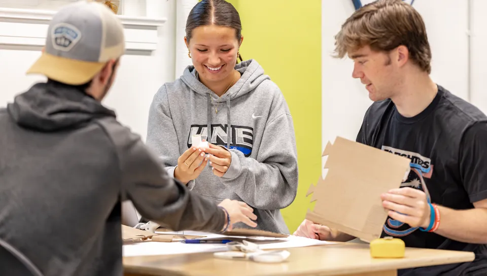 Student in UNE hoodie examining small white device while collaborating with classmates at table.