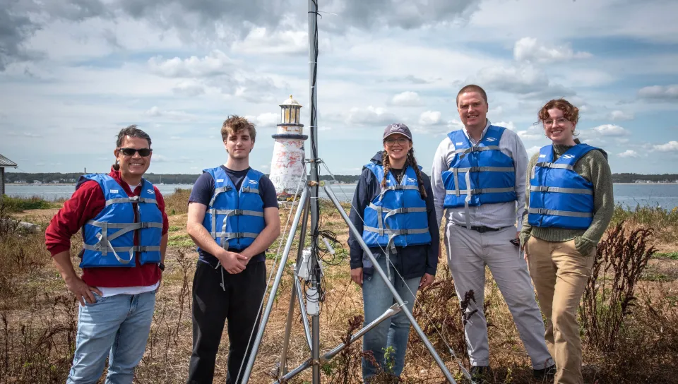 Three Shaw Innovation Fellows with Will Kochtitsky and Charles Tilburg wearing life vests pose with their seismograph equipment and weather station at Ram Island, with a lighthouse and coastal waters visible in the background