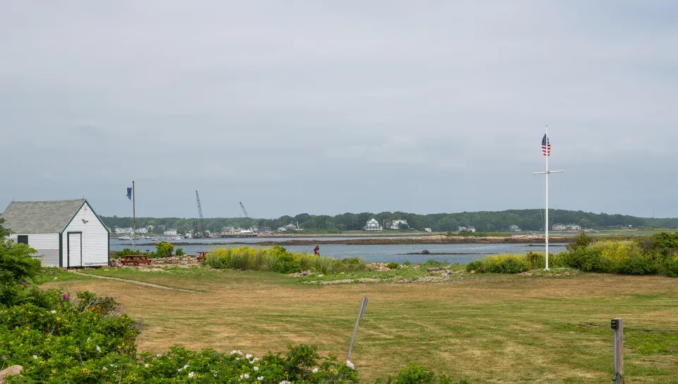 Coastal Maine research station with white outbuilding, grassy field, rocky shoreline, and harbor with distant buildings visible across the water under overcast skies.