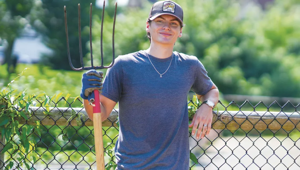 Luke Jenkins stands with a pitchfork at an urban community garden in Portland with a church visible in the background.