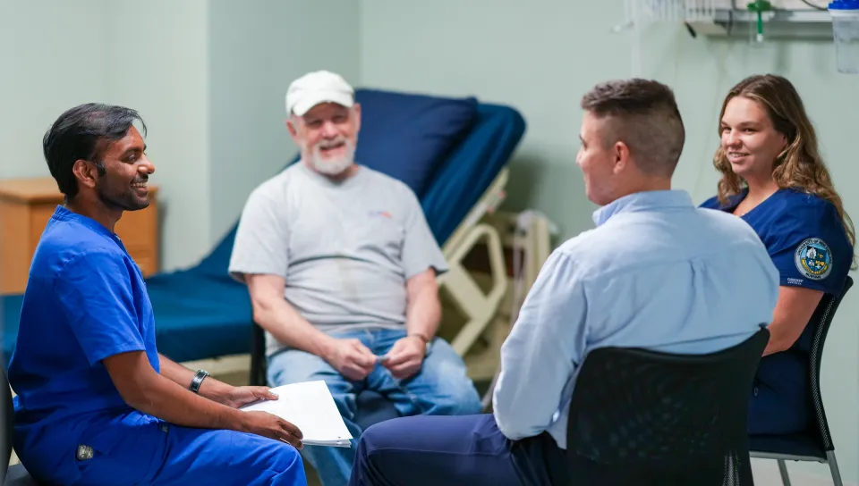 Varun Kota, a College of Osteopathic Medicine student, holds a clipboard while meeting with Michael, a patient actor, alongside Andrii Sokolov and Alexa Lanteri in a clinical simulation room.
