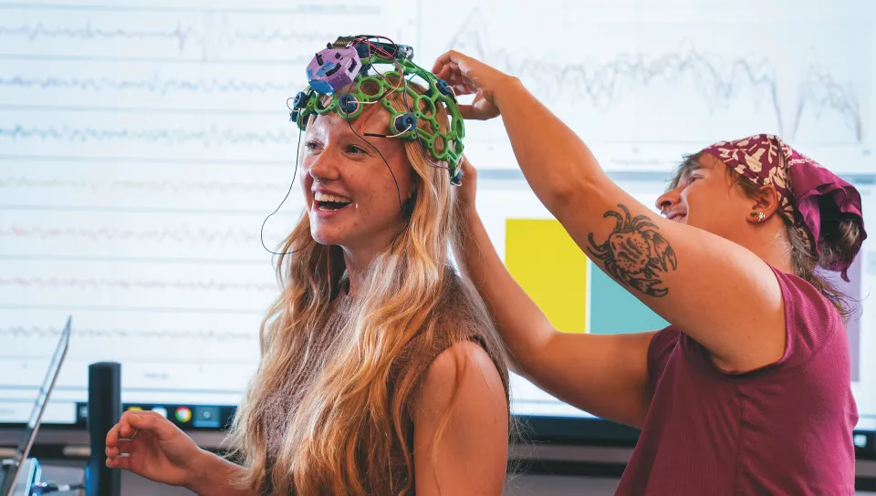 Milo Lypps adjusts a colorful wired skull caps on Jessica Howard's head in a laboratory or classroom setting.