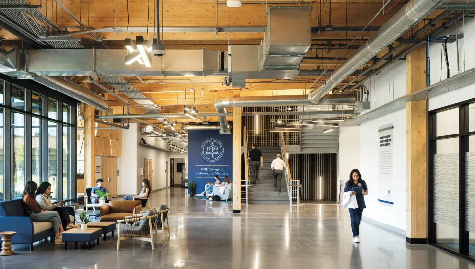Modern UNE College of Osteopathic Medicine building interior with exposed wooden beams, polished concrete floors, central staircase, and students in a contemporary lounge area with natural light from floor-to-ceiling windows.