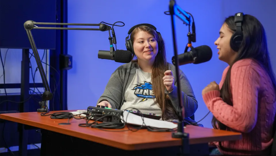 Two students wearing headphones record a podcast in a studio with professional microphones and blue accent lighting.