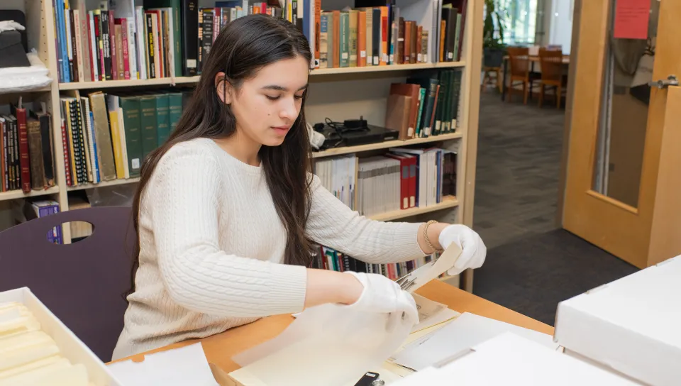 A history student wearing white conservation gloves carefully handles archival documents at a library research table surrounded by reference materials.
