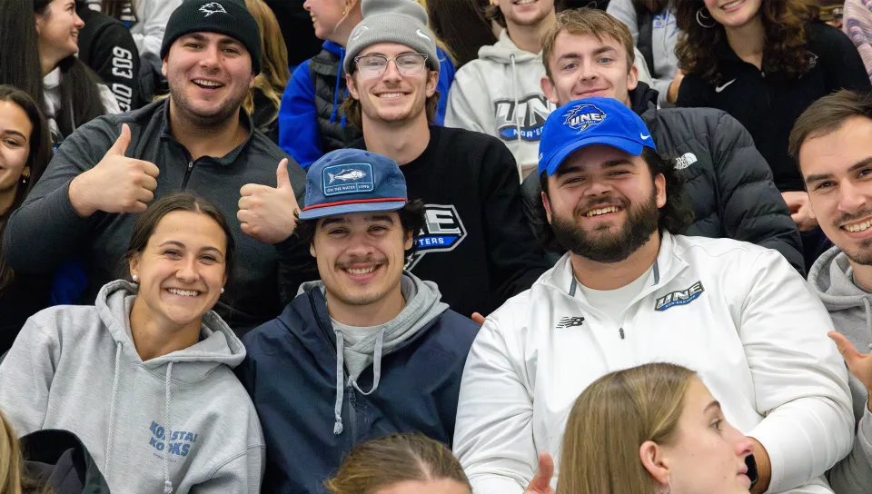 Students gather in bleacher seating at a campus event, smiling and gesturing for the camera.