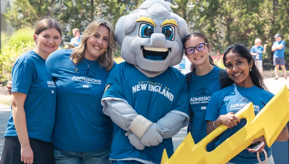 Four admissions student ambassadors pose with the U N E mascot holding a large yellow lightning bolt prop.