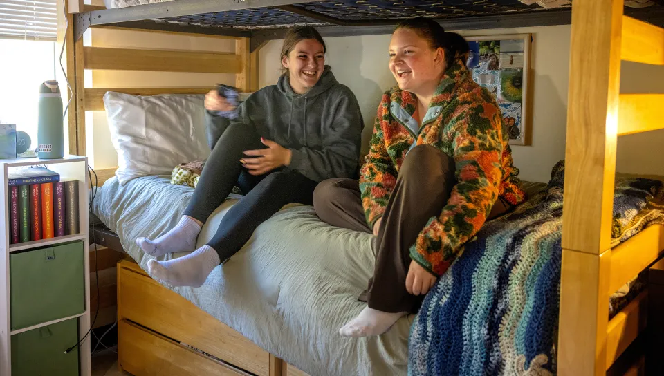 Two roommates laugh together while sitting on beds in their dorm room with personal items and books visible.
