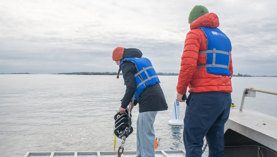 Two marine science students in life jackets deploy sampling equipment from a research vessel into coastal waters.