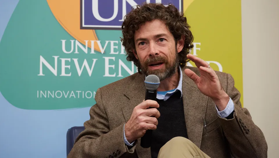 John Waterman holds microphone and gestures while presenting at a President's Forum with branded backdrop.