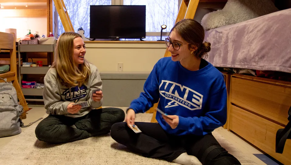 Two students sit cross-legged on a dorm room floor playing cards, one wearing a gray UNE sweatshirt and the other wearing a blue UNE Nor'easters sweatshirt.