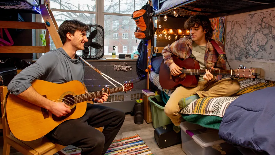 Two students play acoustic guitars together in a dorm room with lofted beds, string lights, decorative tapestries, and a large window overlooking campus buildings.
