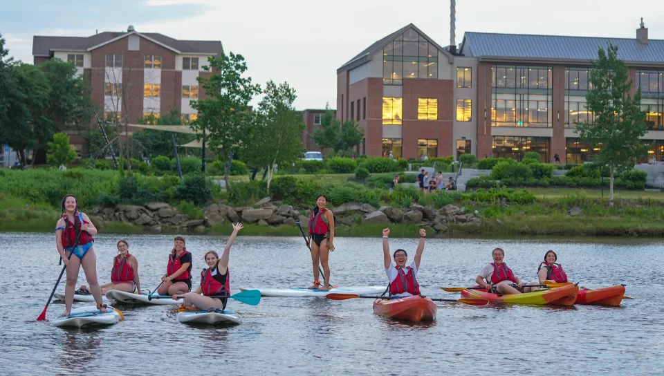 A group of students wearing red life jackets paddle kayaks and stand on paddleboards on the river alongside UNE's campus, cheering and smiling as the campus buildings glow with warm light in the early evening background.