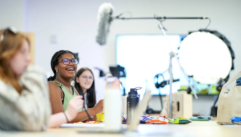 A student laughs during a session in a media production lab, surrounded by professional lighting equipment, a boom microphone, and cameras. Snacks and water bottles are spread across the table, with other students visible in the background.