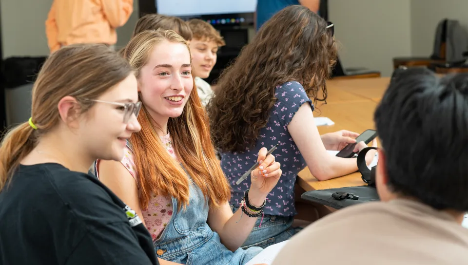 A group of students engaged in lively conversation around a classroom table, with one student smiling and gesturing with a pen. A presentation screen is visible in the background.