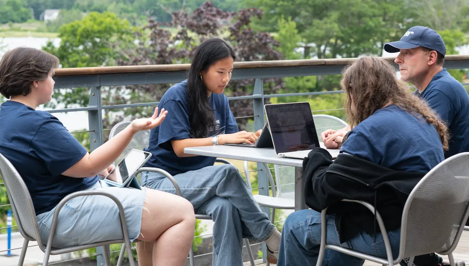 Four students collaborate around a metal table on an outdoor deck, working on laptops and a tablet. Lush green trees and a glimpse of water are visible in the background.