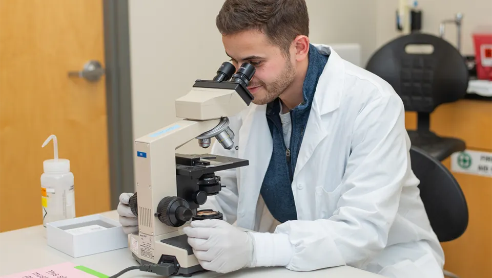  A student in a lab coat and gloves examines a slide under a microscope in a biomedical laboratory.