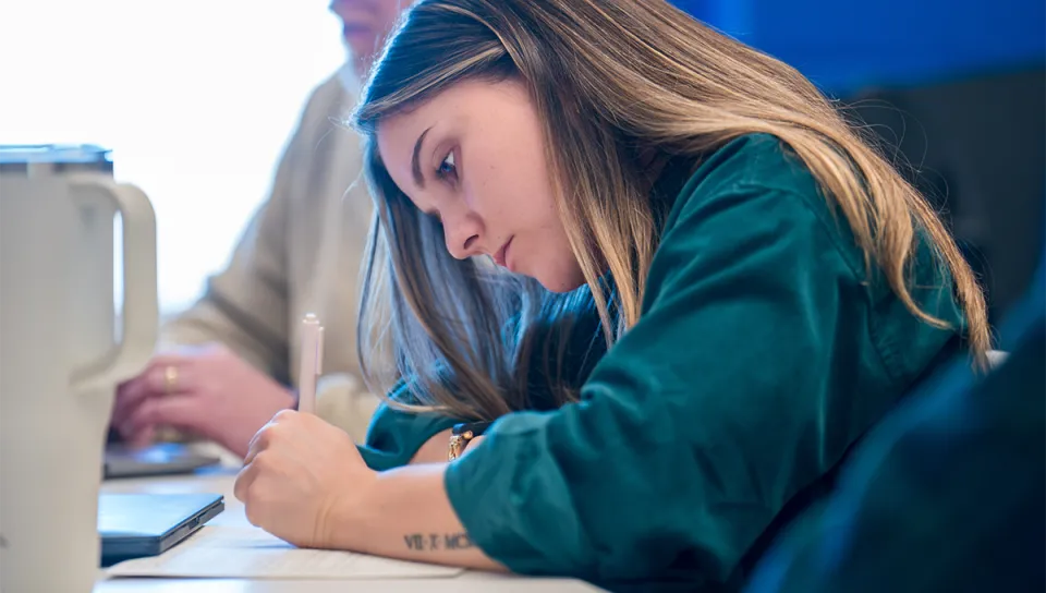 A student writes notes by hand during class, with a laptop and phone on the desk beside her.