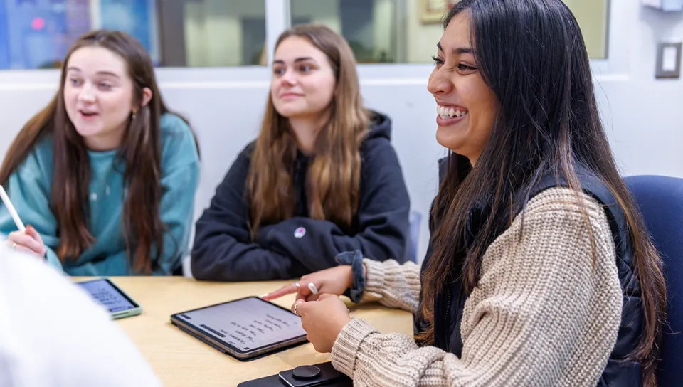Three students sit together at a table engaged in discussion, with tablets and phones in front of them. The student in the foreground smiles broadly while the others listen and react.