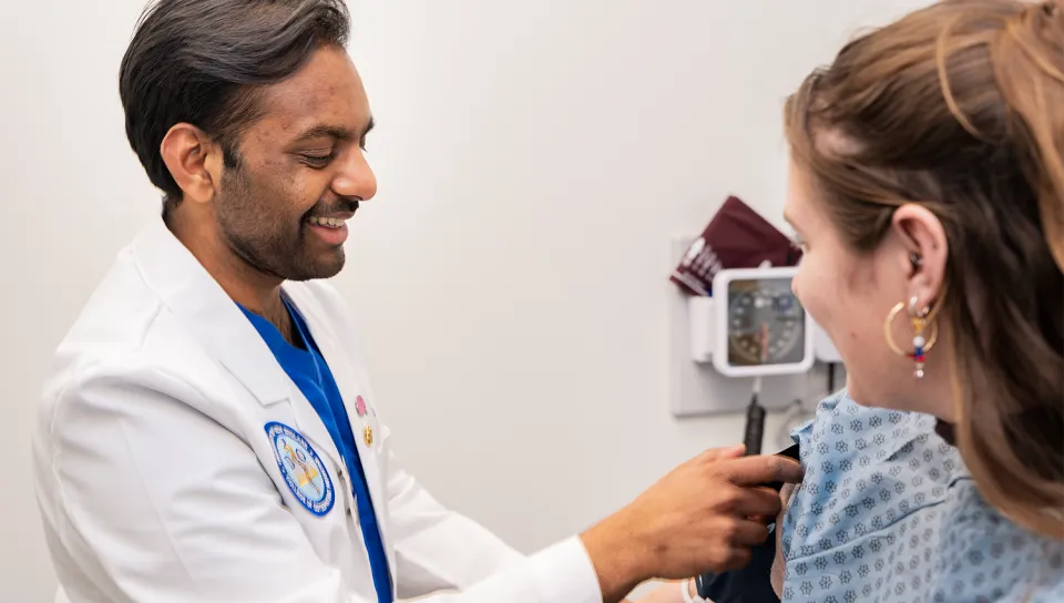 A smiling UNE College of Osteopathic Medicine student in a white coat uses a stethoscope to examine a standardized patient in a clinical practice center exam room.