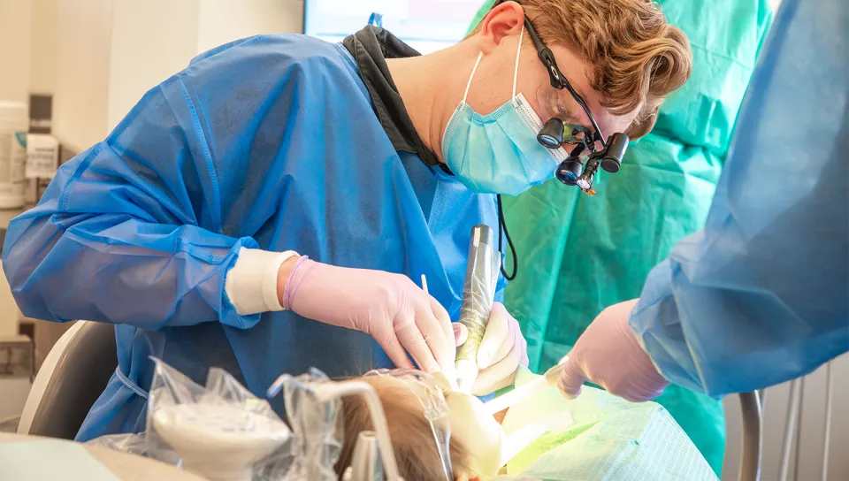 A UNE dental student wearing loupes, a surgical mask, and gloves performs a dental procedure on a patient with the assistance of a second student in a clinical setting.