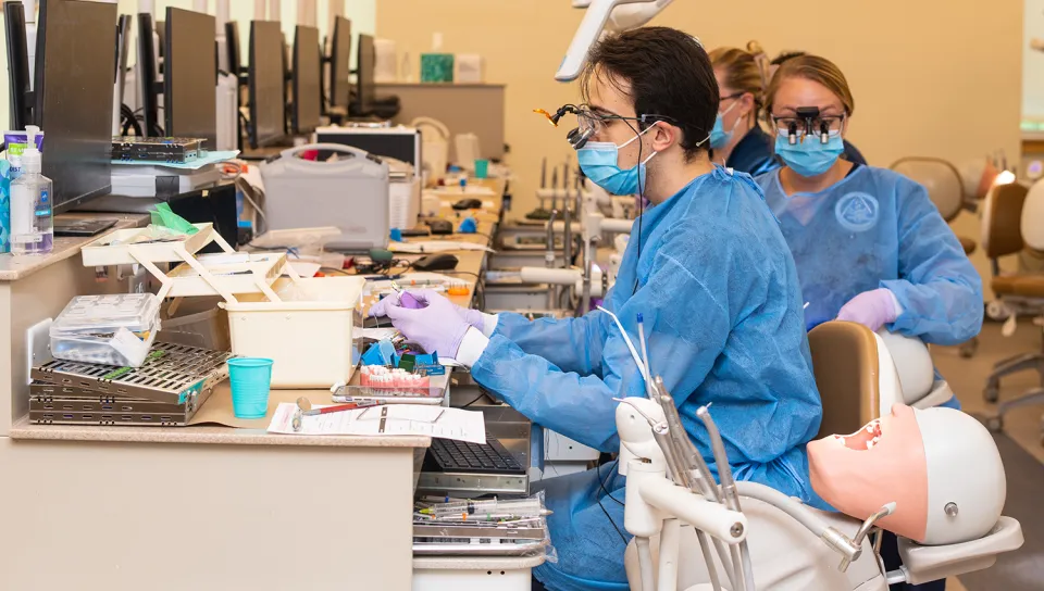 Dental students in blue gowns, masks, and loupes practice procedures on a manikin in a dental skills lab, with instrument trays and workstations visible in the background.