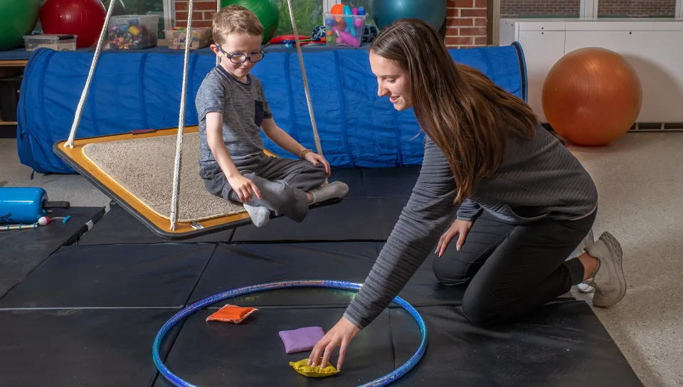 A UNE occupational therapy student sets up a bean bag toss activity inside a hula hoop on a padded mat while a child sits on a platform swing during a pediatric sensory integration session.