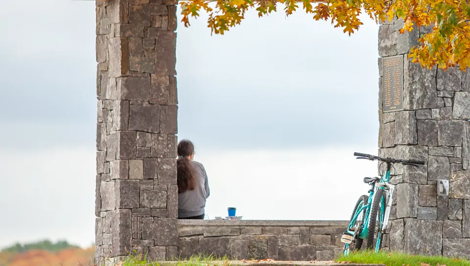A student sits quietly at the Jordan Point stone overlook, gazing toward the water, with a bicycle leaning against the wall and fall foliage framing the scene.