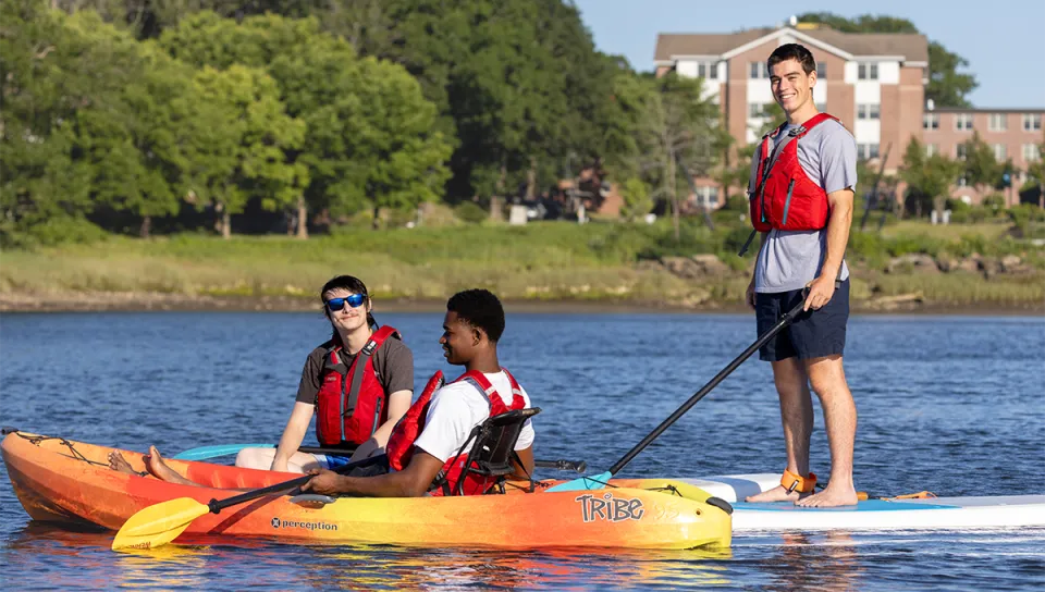 Three UNE students enjoy a sunny day on the water, two kayaking and one stand-up paddleboarding, with campus buildings visible along the tree-lined shore.
