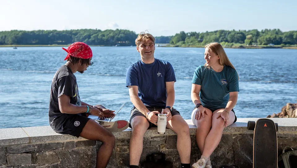 Three U N E students sit by the water on the Biddeford Campus