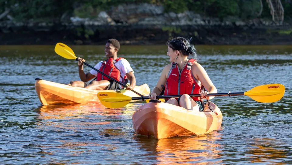  Two students wearing red life vests kayak together on a calm river, smiling as they paddle through the water.