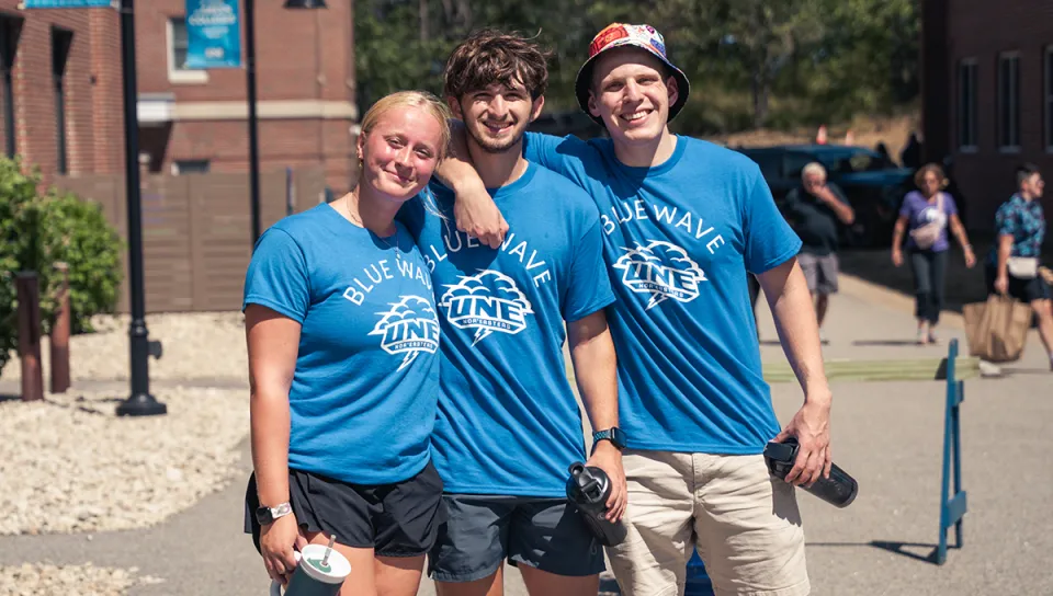 Three students wearing matching blue UNE Blue Wave Nor'easters shirts pose together with arms around each other on campus during an orientation event.