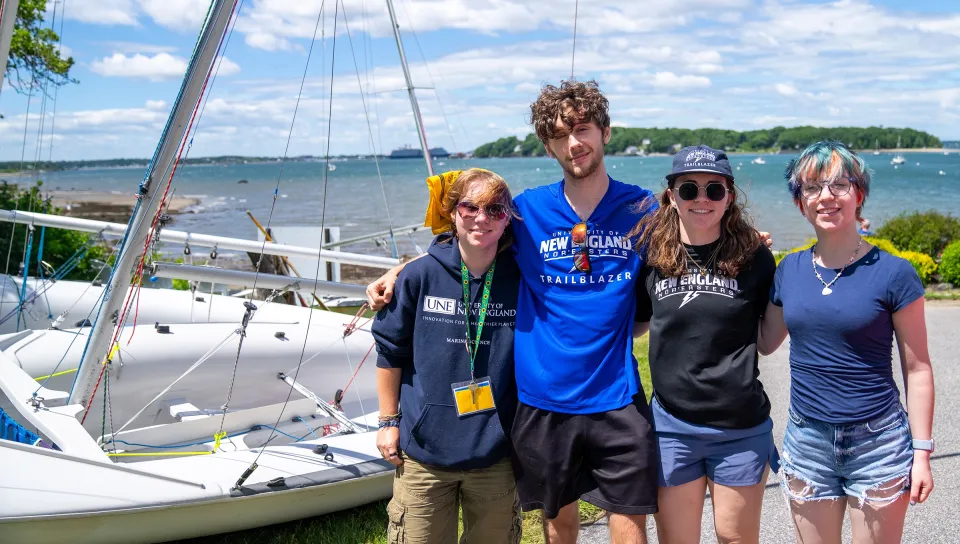 Four UNE Trailblazer students pose together near a sailboat at East End Beach, with Casco Bay and Portland's skyline visible in the background.