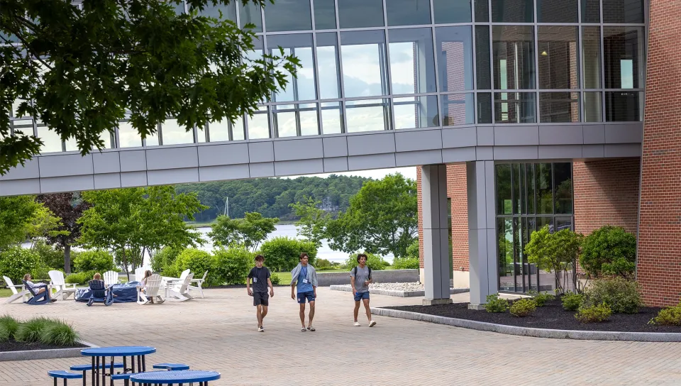 Three students wearing lanyards walk across a sunny brick courtyard beneath a glass pedestrian bridge connecting campus buildings overlooking the water.