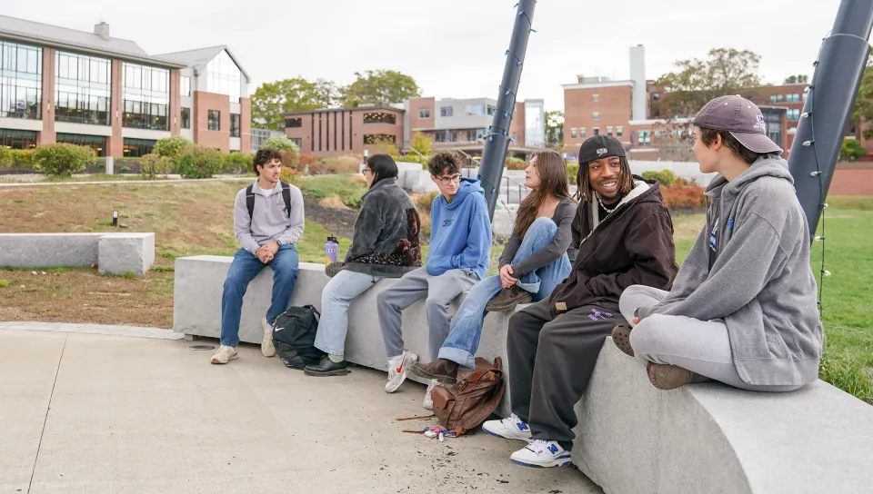 A group of six students sit and chat on concrete benches outdoors on the Biddeford campus, with campus buildings visible in the background.