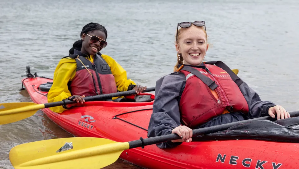 Two students smile from red kayaks on Casco Bay, wearing life jackets and holding paddles.