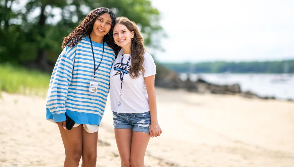 Two students wearing U N E lanyards lean together and smile on a sandy beach with the shoreline and trees blurred behind them.