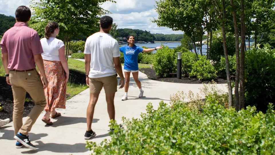 Students walk along a tree-lined path during a Biddeford Campus tour with waterfront views in the background.